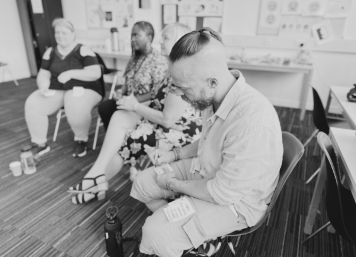 Black and white photo of a white man sitting writing on a post it, three women are in the background doing the same